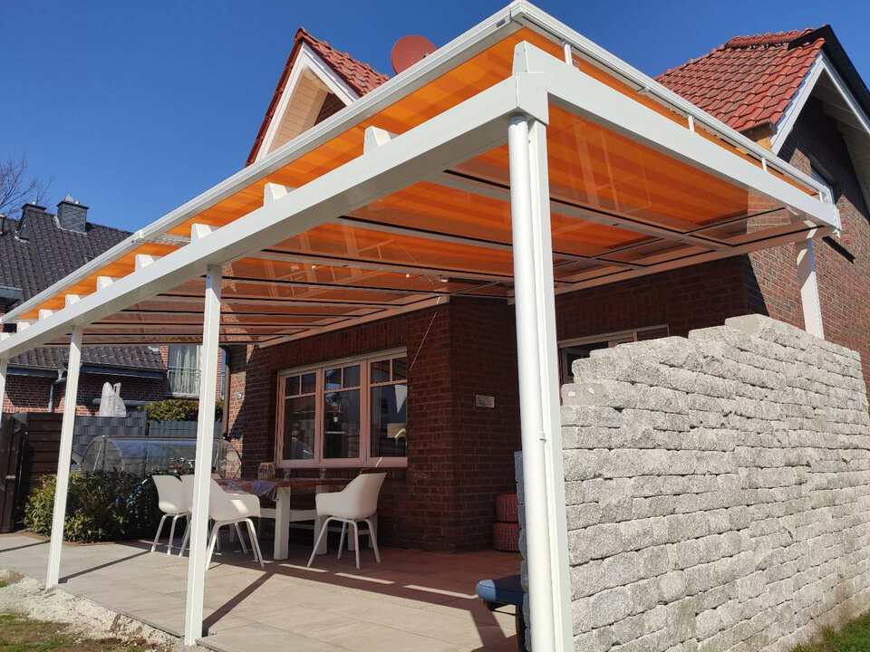 Eine weiße Terrassenüberdachung mit einer integrierten orangefarbenen Aufglasmarkise ist an einem roten Backsteinhaus montiert. Die Überdachung erstreckt sich über eine Terrasse, auf der weiße Gartenstühle um einen Tisch angeordnet sind. Rechts neben der Überdachung befindet sich eine halbhohe Steinmauer.