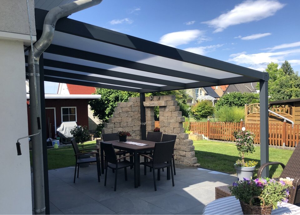 Moderne Terrassenüberdachung mit robusten Aluminiumrahmen und transparenten Polycarbonatplatten. Darunter befindet sich ein Esstisch mit Stühlen, umgeben von einer gepflegten Gartenlandschaft mit Zäunen, Pflanzen und einer dekorativen Steinmauer im Hintergrund.